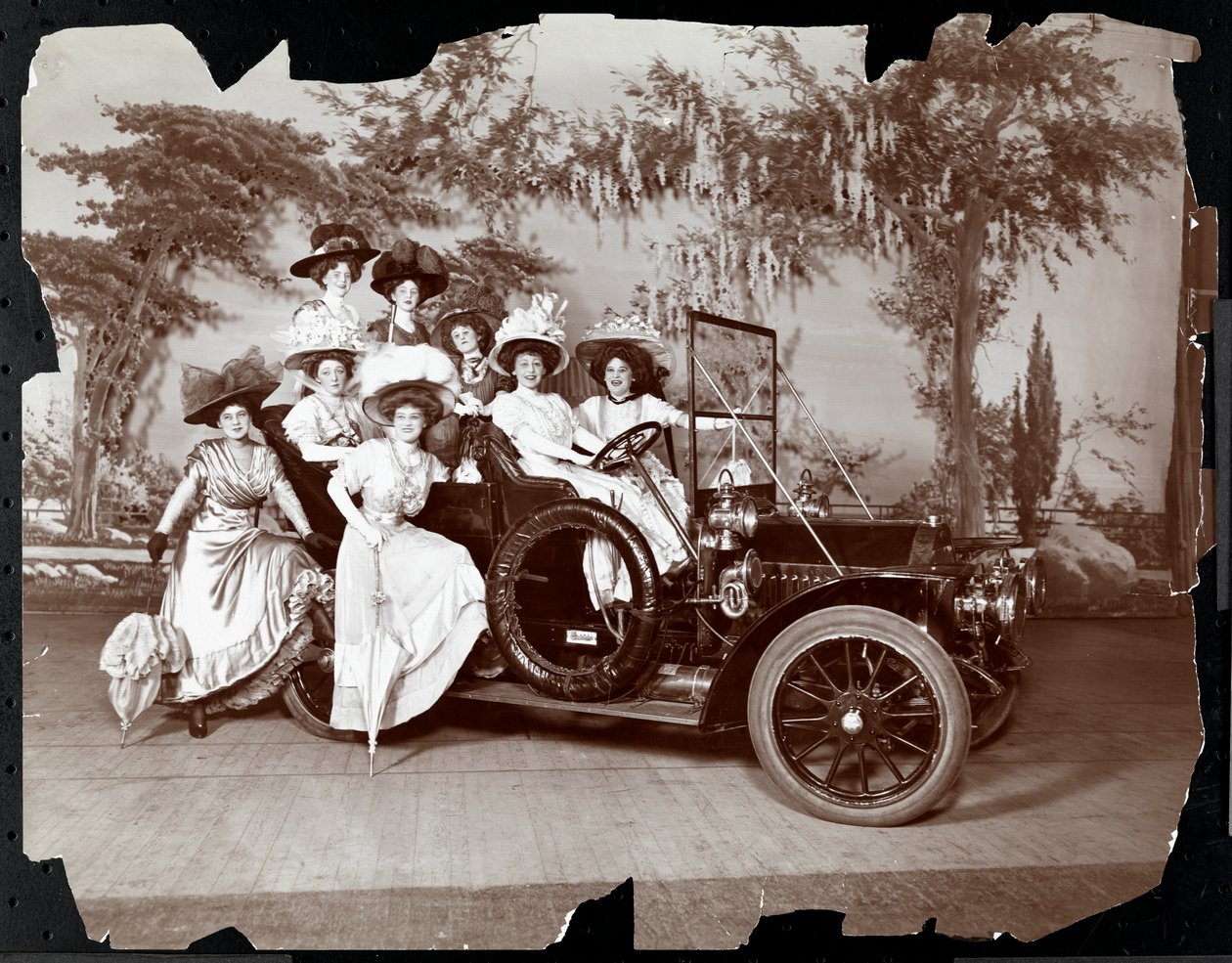 Eight Costumed Women Posed in and Around an Automobile on a Stage with a Landscape Backdrop, Presumably in the Performance of 